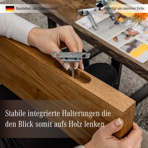 May include: Close-up of a wooden shelf with a metal bracket being installed. The shelf is made of solid oak and has a natural wood finish. The text "Stabile integrierte Halterungen die den Blick somit aufs Holz lenken" is visible on the shelf.