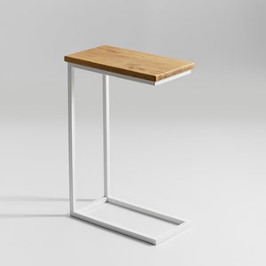 Puede incluir: Una mesa auxiliar moderna con una tapa rectangular de madera clara. La mesa tiene una estructura metálica blanca minimalista. El diseño tiene forma de C, con la tapa que sobresale del marco. La mesa mide aproximadamente 60 cm de largo.