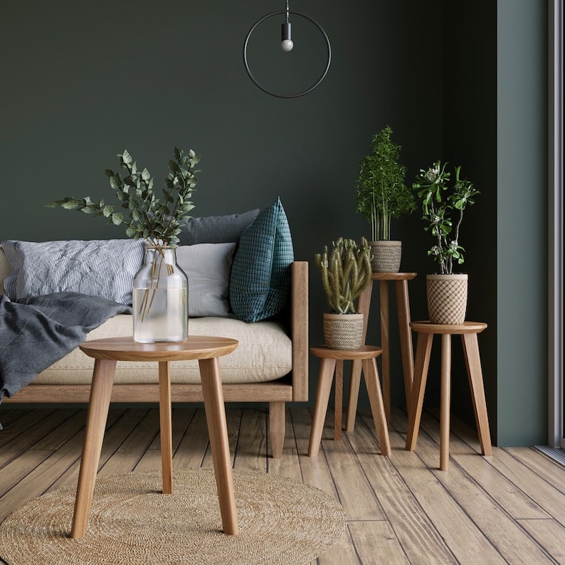 May include: A living room with a couch, three wooden side tables, and a variety of plants in pots. The walls are a dark green, and the floor is a light wood.