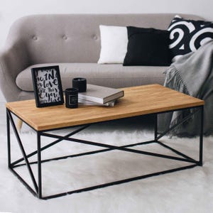 Op de afbeelding: Een houten salontafel met een zwart metalen frame. Het frame heeft een geometrisch ontwerp met kruisende lijnen. De tafel staat op een wit tapijt voor een grijze bank.