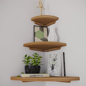 May include: Three wooden corner shelves with rounded edges. The top shelf has a wooden figurine, the middle shelf has a framed geometric print and a white ceramic vase, and the bottom shelf has a potted succulent, a book titled "DONNA TARTT" and a stack of books with the title "A CONFESSION" visible.