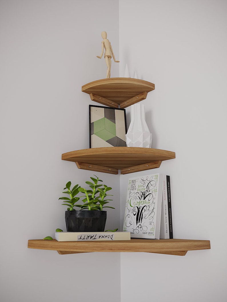 May include: Three wooden corner shelves with rounded edges. The top shelf has a wooden figurine, the middle shelf has a framed geometric print and a white ceramic vase, and the bottom shelf has a potted succulent, a book titled "DONNA TARTT" and a stack of books with the title "A CONFESSION" visible.