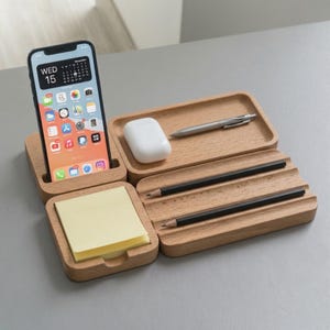 Op de afbeelding: Een bureau-organizer van lichtgekleurd hout met een verticale gleuf voor een smartphone, een vierkant vak met gele memoblaadjes, een rechthoekig bakje met witte draadloze oordopjes en een zilveren pen, en twee gegroefde sleuven.
