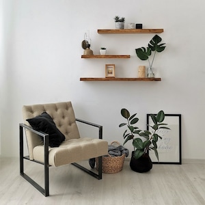 May include: A beige armchair with a black pillow sits in front of a white wall with three wooden shelves. The shelves are decorated with plants, a small picture frame, and candles. A black and white framed print with the text "Let's stay home" is on the floor.
