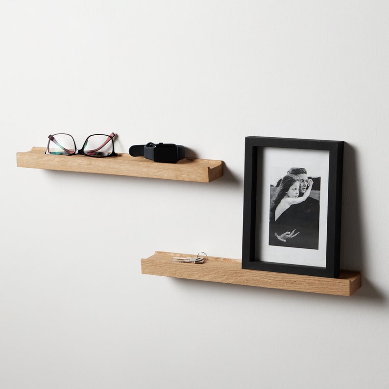 Puede incluir: Dos estantes flotantes de madera montados en una pared blanca. El estante superior tiene un par de gafas y un reloj negro. El estante inferior tiene un marco de fotos con una foto en blanco y negro de una pareja y un juego de llaves.