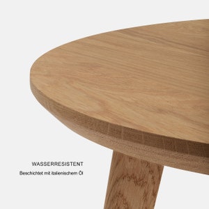 May include: A close-up of a light brown wooden table with a rounded oval top and three legs. The table is water-resistant and coated with Italian oil. The text "WASSERRESISTENT" and "Beschichtet mit italienischem &Ouml;l" is visible on the table.