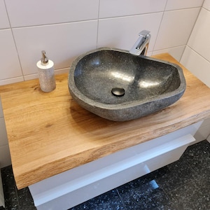 Könnte beinhalten: Ein dunkelgraues Steinklobecken mit abgerundeter Form sitzt auf einer hellbraunen Holztheke. Das Waschbecken hat einen verchromten Wasserhahn und einen kleinen schwarzen Abflussstopfen. Die Arbeitsplatte ist an einem weißen Schrank befestigt.