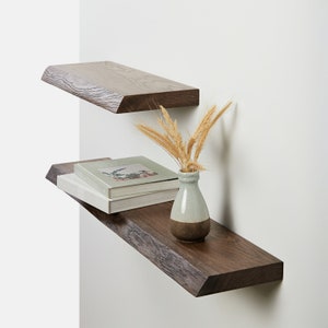 Könnte beinhalten: Zwei Holzregale an einer weißen Wand montiert. Das obere Regal ist kürzer und das untere Regal ist länger. Das untere Regal hat einen Stapel Bücher und eine kleine Vase mit getrockneten Weizenhalmen.