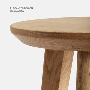 May include: A close-up of a light wood round table with three legs. The table top has a slightly rounded edge. The text "ELEGANTES DESIGN" and "Handgeschliffen" is visible in the top left corner of the image.