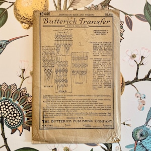 May include: Vintage Butterick Transfer pattern envelope. The tan envelope features printed text and illustrations for needlework smocking designs. The text includes instructions and measurements in inches. The background is a colorful floral wallpaper.