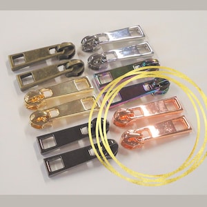 May include: An assortment of metal zipper pulls in various colors, including bronze, silver, gold, black, iridescent, and rose gold. Each pull has a rectangular tab with a square hole and a rounded zipper head. These are ideal for sewing and crafting projects.
