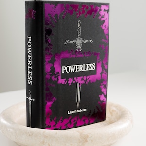 May include: A black hardcover book with a pink and silver foil design. The title "POWERLESS" is printed in silver on the front cover, with the author's name "Lauren Roberts" printed below. A silver sword design is featured on the front and spine.