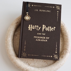 May include: A hardcover book titled "Harry Potter and the Prisoner of Azkaban" by J.K. Rowling. The book is dark brown with gold lettering and decorative star accents. A gold charm bookmark is attached. The book rests on a light-colored, round tray.
