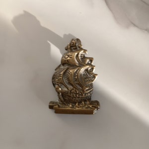 May include: An antique brass ship figurine with detailed carvings. The ship features multiple sails and is mounted on a rectangular base. The brass has a warm, aged patina. The figurine is set against a white marble background.