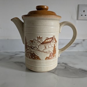 May include: A beige ceramic coffee pot with a brown lid and handle. The pot features a brown landscape design with a house and trees. The pot is sitting on a white surface.