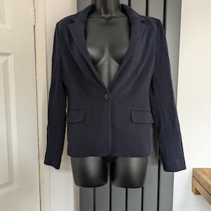May include: A navy blue blazer with a single-button fastening, two front pockets, and a notched lapel. The blazer is displayed on a black mannequin. The garment is made of a soft fabric.