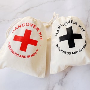 Peut inclure: Deux sacs en coton écrus avec cordon de serrage, imprimés avec "Hangover Kit" et une croix. L'un a une croix rouge, l'autre une croix noire. Le texte "In Sickness and In Health" est également imprimé. Sur une surface en marbre blanc.