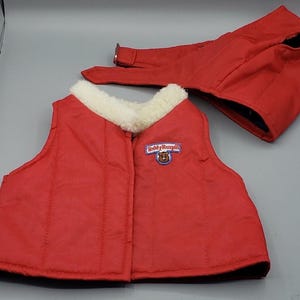 May include: A red vest with a white faux fur collar and a Teddy Ruxpin patch on the chest, accompanied by a matching red hat with a buckle strap. The vest features vertical stitching and the Teddy Ruxpin logo is embroidered in blue and white.