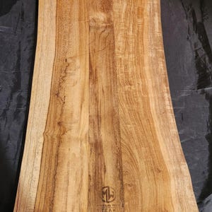 Puede incluir: Una tabla de cortar rectangular de madera con borde natural. La tabla es de madera de color marrón claro con vetas visibles y un logotipo que dice "Mountain Wood Teak".