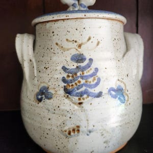 May include: A ceramic jar with a lid, featuring a speckled white glaze and blue floral designs. The jar has two handles and a lid with two decorative knobs. The overall design is rustic and handmade, with a focus on natural elements.
