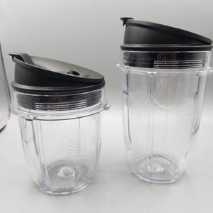 May include: Two clear plastic blender cups with black lids. The cups are different sizes, with the larger one on the right. The lids have a flip-top design. The brand name "Ninja" is printed on the side of the cups.