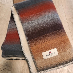 Puede incluir: Una manta doblada con un degradado de colores, incluyendo tonos de rojo, gris, marrón y beige. La manta tiene un borde blanco y un borde cosido. Una etiqueta rectangular con la palabra "WOOLRICH" es visible.
