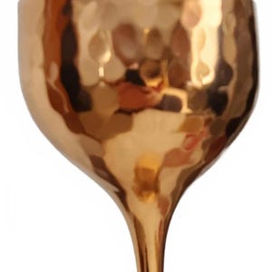 May include: A gold-colored goblet with a hammered texture. The goblet has a wide bowl and a slender stem. The surface reflects light, creating highlights and shadows. The overall design is elegant and decorative.