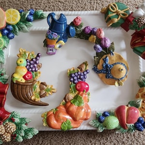 May include: A white ceramic serving tray with a variety of colorful ceramic ornaments. The ornaments include a cornucopia filled with fruit, a watering can with flowers, a pineapple, a pumpkin, a hat, and two wrapped presents.