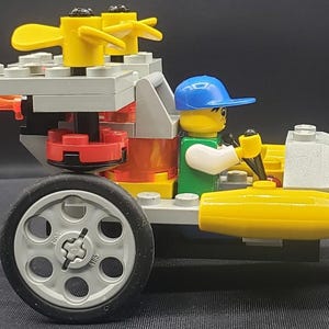 Puede incluir: Un vehículo de juguete Lego con un conductor de minifigura que lleva una gorra azul. El vehículo es principalmente amarillo, gris y rojo, con detalles de llamas naranjas. Tiene una rueda trasera grande y una rueda delantera más pequeña.