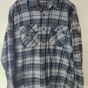 May include: A long-sleeved, button-down flannel shirt in a plaid pattern of black, grey, and light blue. The shirt has two chest pockets and a small patch on the sleeve. The brand name "HOWITZER" is visible on the collar and pockets.