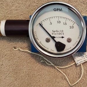 May include: A circular flow meter gauge with a white face, black needle, and silver metal rim. The gauge displays GPM, SpGr=1.0, and Vis=1.0CS. It is mounted on a blue square base with dark brown cylindrical extensions and a brass barbed fitting.