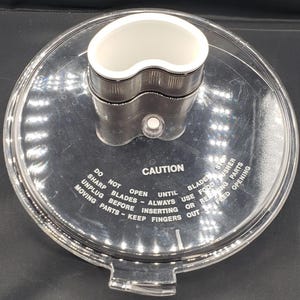 May include: A clear plastic food processor lid with a white and silver central component. The lid features a warning label with the word "CAUTION" and other safety instructions. The lid is designed for a food processor.