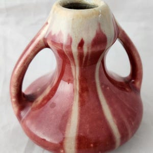 Peut inclure: Un petit vase en céramique rayé rouge et blanc avec deux anses. Le vase a un corps arrondi et un col étroit.