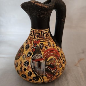 May include: A small, black ceramic pitcher with a rooster design. The rooster is painted in red, yellow, and black on a cream background. The pitcher has a black handle and a geometric design around the top.