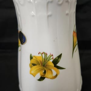 May include: White ceramic vase with a flared top and floral design. The vase features yellow lilies with green leaves and accents of blue and orange. The vase has a decorative embossed pattern.