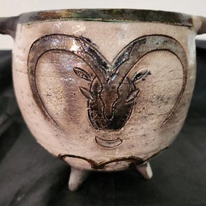 May include: A handcrafted ceramic bowl with a ram's head design. The bowl is a light beige color with a dark brown rim and accents. It has three small feet and two handles. The ram's head is etched into the bowl's surface.