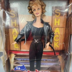 Può includere: Una bambola Barbie con giacca in similpelle nera e tuta nera, confezionata in una scatola con l'etichetta Collector Edition e Grease 25 years. La bambola ha capelli biondi ricci e orecchini a cerchio dorati davanti a uno sfondo Shake Shack.