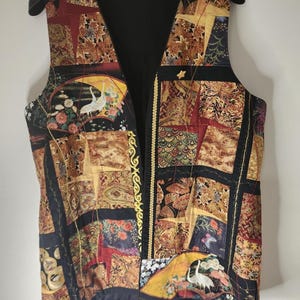Op de afbeelding: Een mouwloos vest met een patchworkontwerp van diverse stoffen in zwart, goud, rood en geel. Het patroon bevat afbeeldingen van witte kraanvogels, bloemmotieven en geometrische vormen. De opening aan de voorzijde is afgezet met een gouden sierrand.
