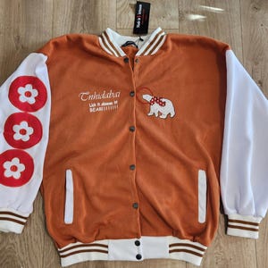 May include: An orange varsity jacket with white sleeves and accents. The jacket features a floral design on the left sleeve and a bear graphic with text on the front. The jacket has a button-up closure and two front pockets.