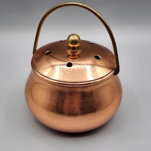 Könnte beinhalten: Ein kupferfarbener, runder Topf mit Deckel und Griff. Der Deckel hat kleine Löcher und einen goldfarbenen Knopf. Der Griff ist ebenfalls goldfarben und wölbt sich über den Topf. Der Topf hat eine glänzende Oberfläche.