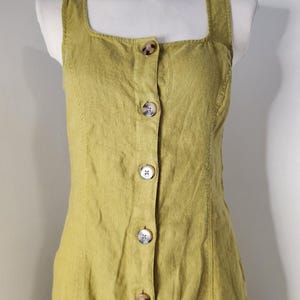 Op de afbeelding: Een mouwloze, olijfgroene jurk met een vierkante halslijn en een knoopsluiting aan de voorkant. De jurk is gemaakt van een textuurstof en heeft grote, ronde knopen. De jurk wordt op een mannequin getoond.
