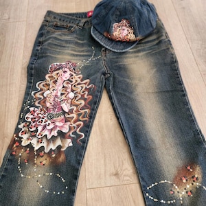 May include: Blue denim jeans and matching cap. The jeans feature a hand-painted design of a woman with long, wavy blonde hair, wearing a floral and leopard print outfit. The cap has a similar design. The jeans have a faded wash.