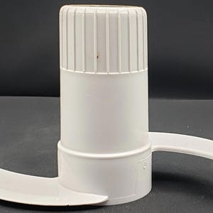 May include: A white food processor blade assembly. The assembly has a cylindrical body with a ridged top and a curved blade extending from the base. The blade is designed for chopping and processing food items.