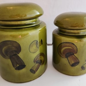 May include: Two olive green ceramic canisters with matching lids. The canisters are decorated with brown and tan mushroom illustrations. The larger canister is approximately 8 inches tall, and the smaller one is about 6 inches tall. The lids are rounded.