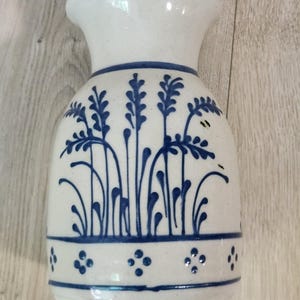 Op de afbeelding: Een witte keramische vaas met een uitlopende rand, versierd met handgeschilderde blauwe botanische patronen met hoge grasachtige stengels en een horizontale band van kleine vierpuntige bloemmotieven nabij de basis.