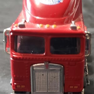 Puede incluir: Un camión semirremolque de juguete rojo con una parrilla plateada y el logotipo de Kenworth. El camión tiene una cabina con dos ventanas y un techo con un logotipo. El camión tiene luces amarillas en el techo y en la parte delantera.