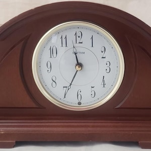 May include: A wooden mantel clock with a dark brown finish. The clock face is white with black numerals and gold-colored hands. The clock has a gold-colored bezel and decorative gold accents.