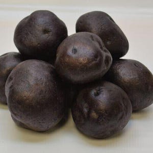Puede incluir: Un montón de patatas moradas oscuras con una textura ligeramente rugosa. Las patatas tienen forma ovalada y varían en tamaño, con un color profundo y rico. La imagen es un primer plano, destacando los detalles del producto.