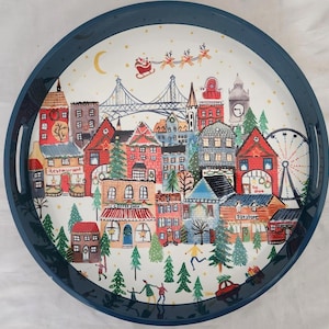 以下が含まれることがあります： 青い縁取りと白い背景を持つ丸いサービングトレイで、お祝いの冬の村のシーンが描かれています。シーンには橋、観覧車、コーヒーショップ、おもちゃ屋、レストランが含まれています。サンタクロースはトナカイに乗って空を飛んでいます。