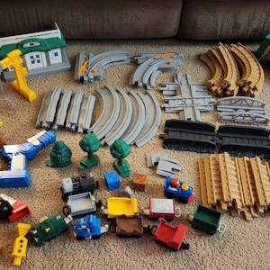 May include: A large collection of plastic toy train set components including various track pieces in gray, tan, and green, a blue crane, a red and white building, a white barn, small train cars, and assorted plastic signs and trees.